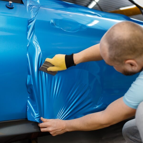 Male mechanic installs protective vinyl foil or film on vehicle door. Worker makes auto detailing. Automobile paint protection, professional tuning
