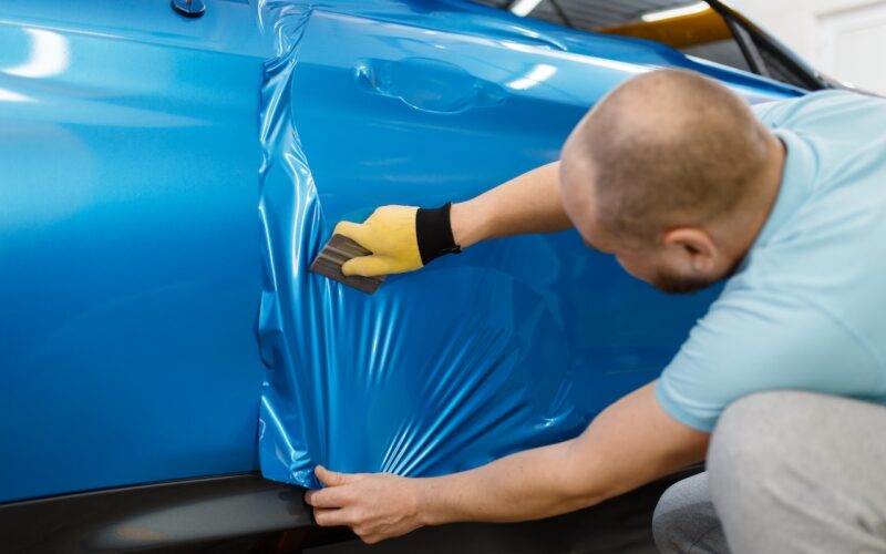 Male mechanic installs protective vinyl foil or film on vehicle door. Worker makes auto detailing. Automobile paint protection, professional tuning